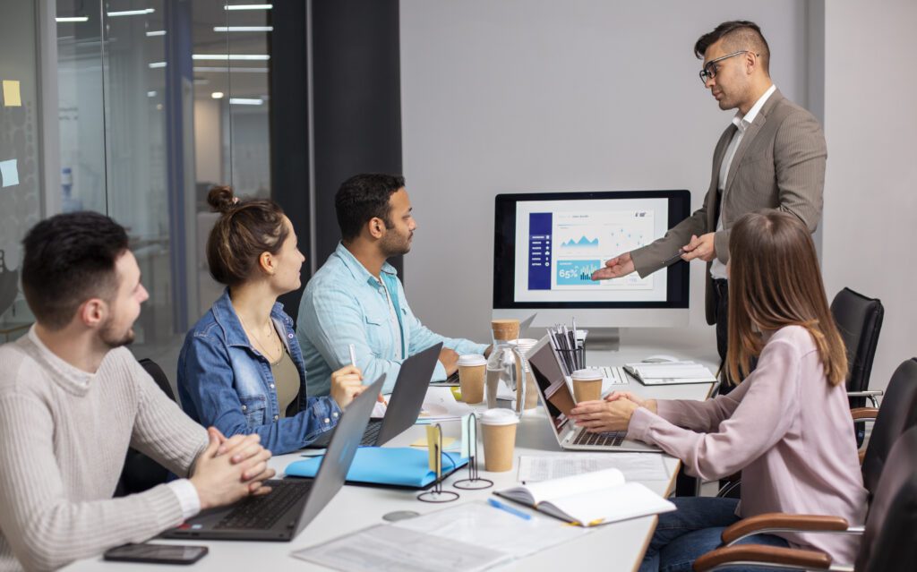 a man standing in front of a computer screen inside a conference room having a meeting with colleagues about Sales Closing Tactics
