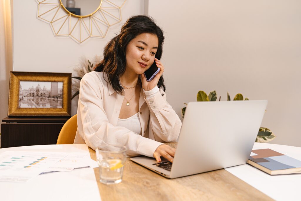 A woman holding a cellphone with her left hand talking to a client while the right hand is typing on the laptop