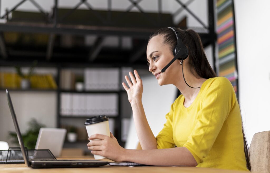 a woman wearing a yellow t-shirt with a headset on her head waving her hand to the computer screen.