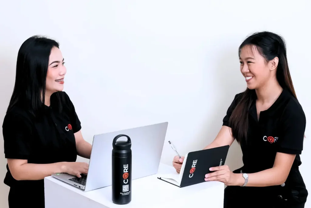 Two women in black shirts collaborate at a table, each using a laptop, discussing clinical trial recruitment challenges.