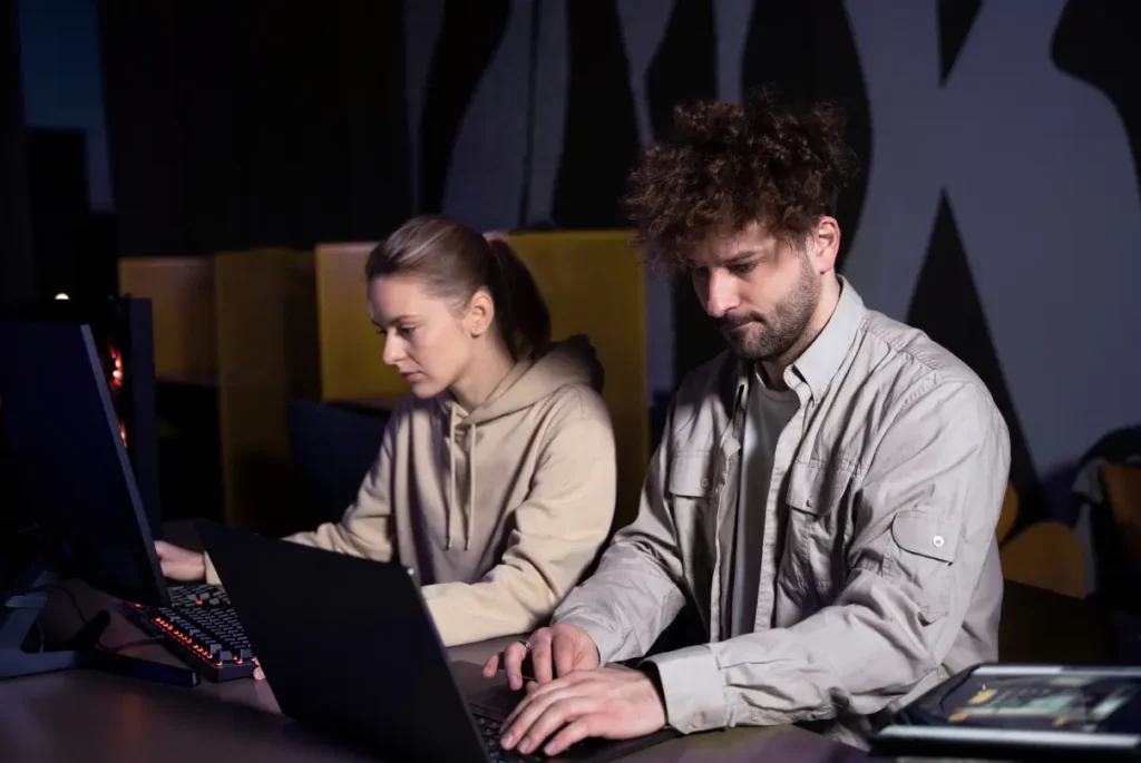 Two individuals focused on their computers in a dimly lit room, emphasizing cybersecurity solutions for business.