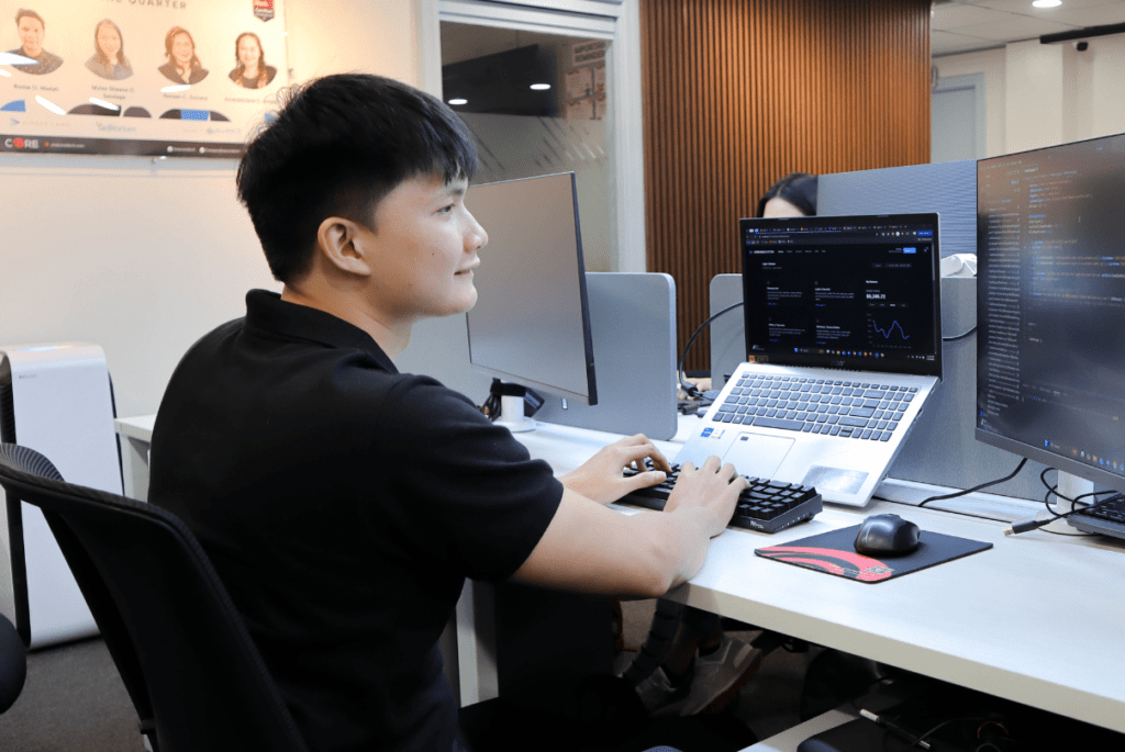 A man seated at a desk with two computers, working on IT services for education.