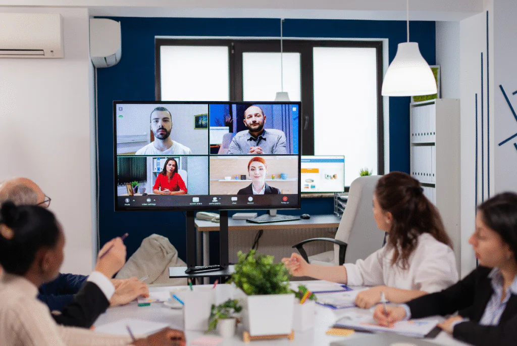 A group of people in a conference room discussing remote marketing team strategies, with a large screen displaying data.