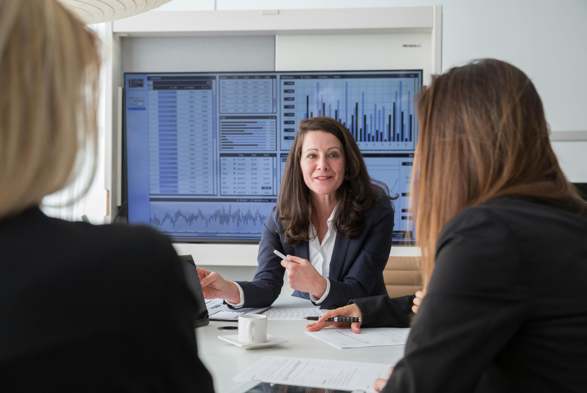 A woman in a business suit discusses outsourcing versus layoffs with two colleagues in a professional setting.