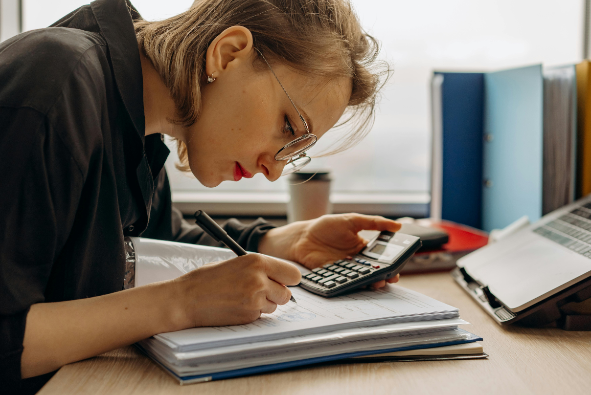 A woman calculates figures on a calculator while taking notes in a notebook, focused on outsourced finance and accounting tasks.