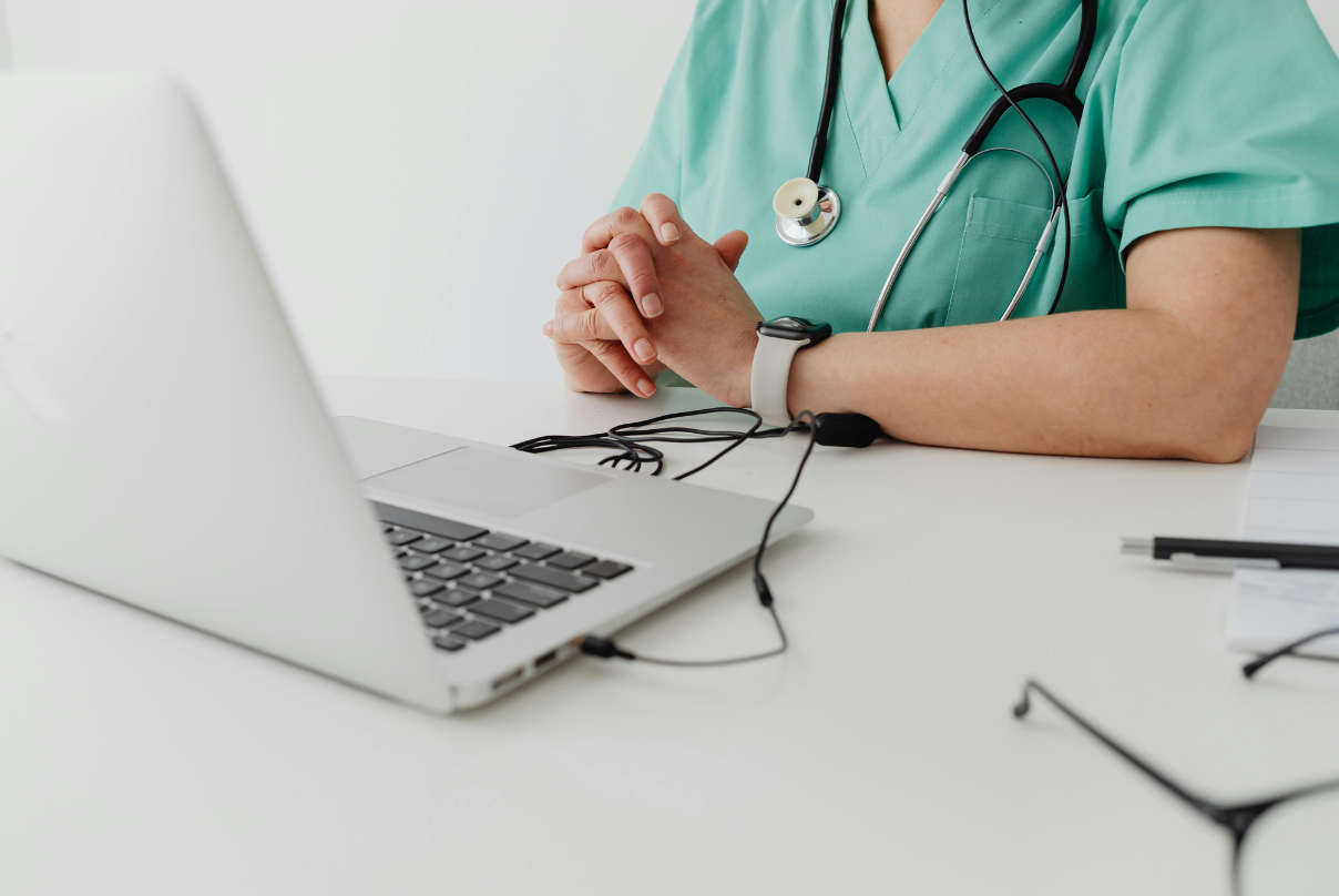 A woman in scrubs sits at a desk with a laptop, engaged in telehealth outsourcing work.