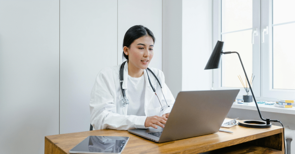 A woman in a white coat sits at a desk with a laptop, engaged in telehealth outsourcing activities.