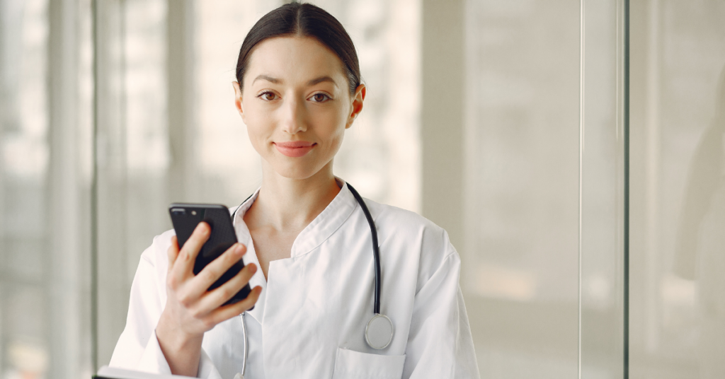 The photo shows a woman in an indoor setting wearing a white long sleeve top and a black stethoscope around her neck while holding a phone
